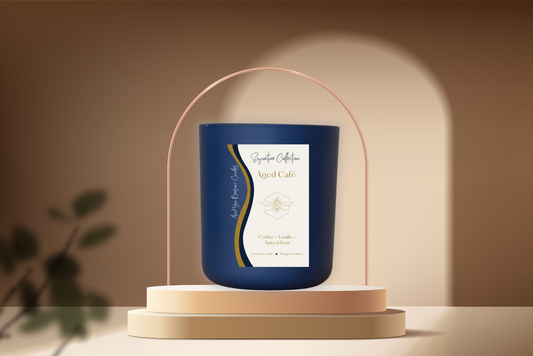 An Aged Café Luxury Beeswax Candle displayed beautifully. The candle is adorned with a label that features the scent name, and its flickering flame casts a cozy glow, adding a touch of warmth to the setting